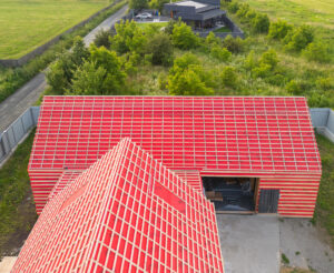 Vedere aeriană a unei case vibrante cu acoperiș roșu, situată în mijlocul unei vegetații luxuriante și a unor câmpuri. Peisajul prezintă un amestec de clădiri moderne și rustice.