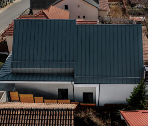 Vedere aeriană a caselor cu acoperiș metalic modern, gri închis. Se văd strada și casele învecinate cu acoperișuri roșii și gri.