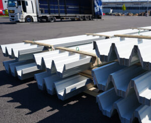 Stive de tablă albă de acoperiș metalică pe paleți de lemn, cu un camion cu laterale deschise în fundal.