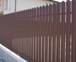 Un gard metalic maro cu panouri verticale se află în fața unei case de culoare crem cu acoperiș înclinat. Scena transmite intimitate și siguranță.