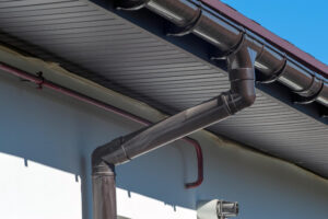 Prim-plan al unui jgheab și al unei burlane metalice de pe acoperișul unei clădiri, sub un cer albastru senin.