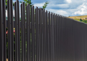 Un șir de stâlpi de gard din metal închis la culoare se întinde în diagonală pe imagine, cu un cer înnorat și câmpuri îndepărtate în fundal, creând o senzație de profunzime.