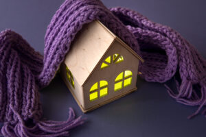 O căsuță din lemn cu ferestre galbene strălucitoare este înfășurată într-o eșarfă violetă confortabilă, emanând căldură și confort pe un fundal neutru.