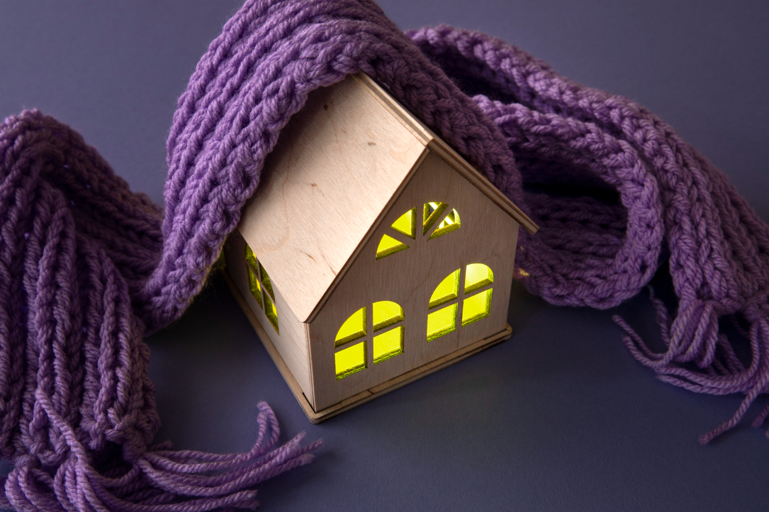 O căsuță din lemn cu ferestre galbene strălucitoare este înfășurată într-o eșarfă violetă confortabilă, emanând căldură și confort pe un fundal neutru.