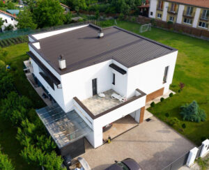 O casă modernă, cubică, de culoare albă, cu acoperiș plat de culoare închisă, o terasă spațioasă în aer liber și o grădină luxuriantă. Cadrul este liniștit și suburban.