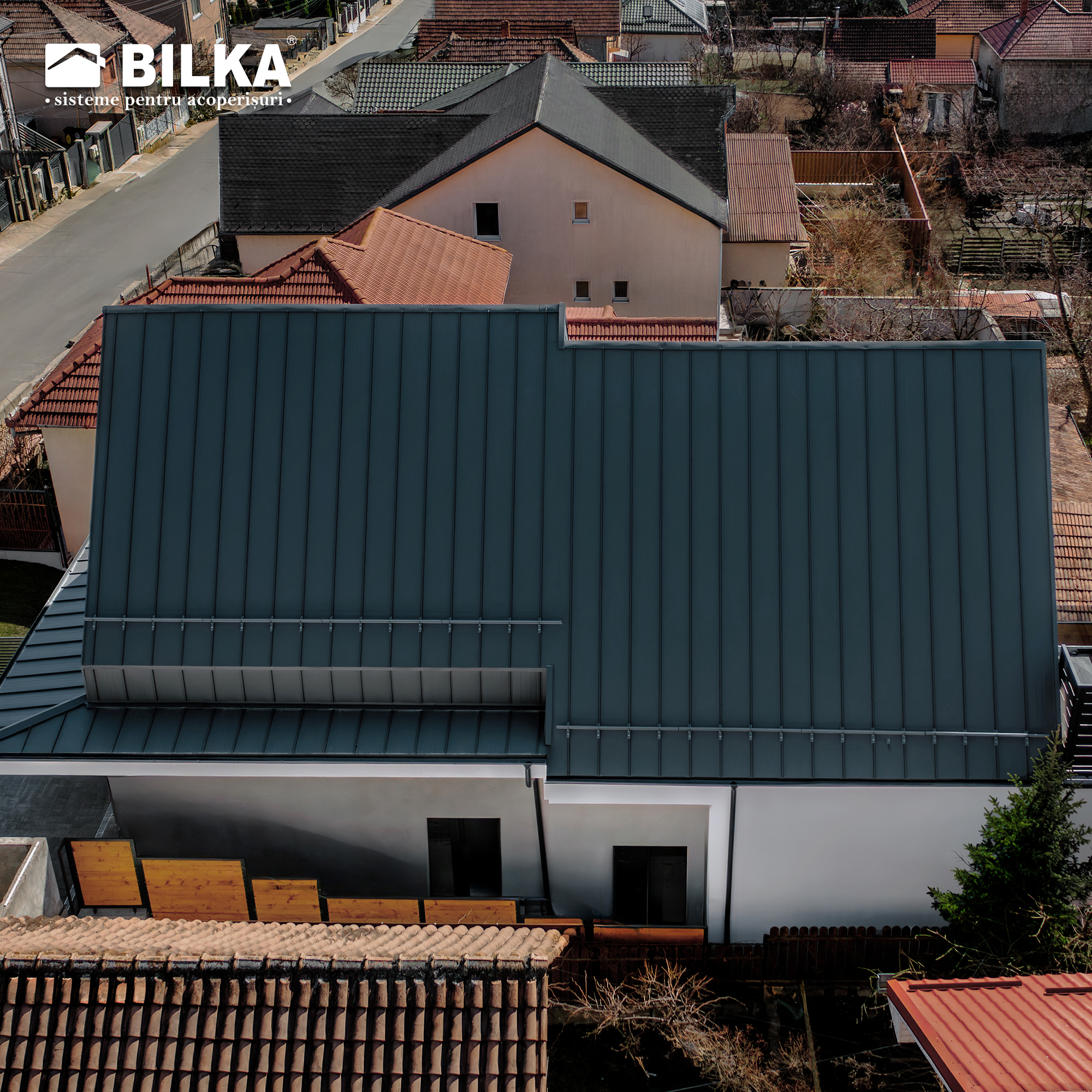 Vedere aeriană a caselor cu acoperiș metalic modern, gri închis. Se văd strada și casele învecinate cu acoperișuri roșii și gri.