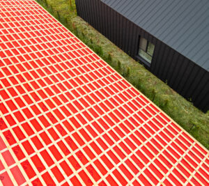 Vedere aeriană a unui acoperiș în carouri roșii, sub care se află o clădire neagră cu o fereastră. Peisajul este înconjurat de iarbă verde și arbuști.