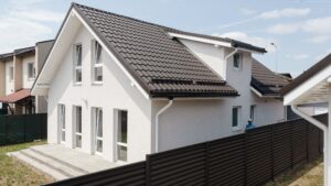 O casă modernă cu două etaje, cu acoperiș din țiglă închisă la culoare și pereți albi. Dispune de ferestre mari și o curte împrejmuită, sub un cer albastru senin, emanând o atmosferă contemporană și primitoare.