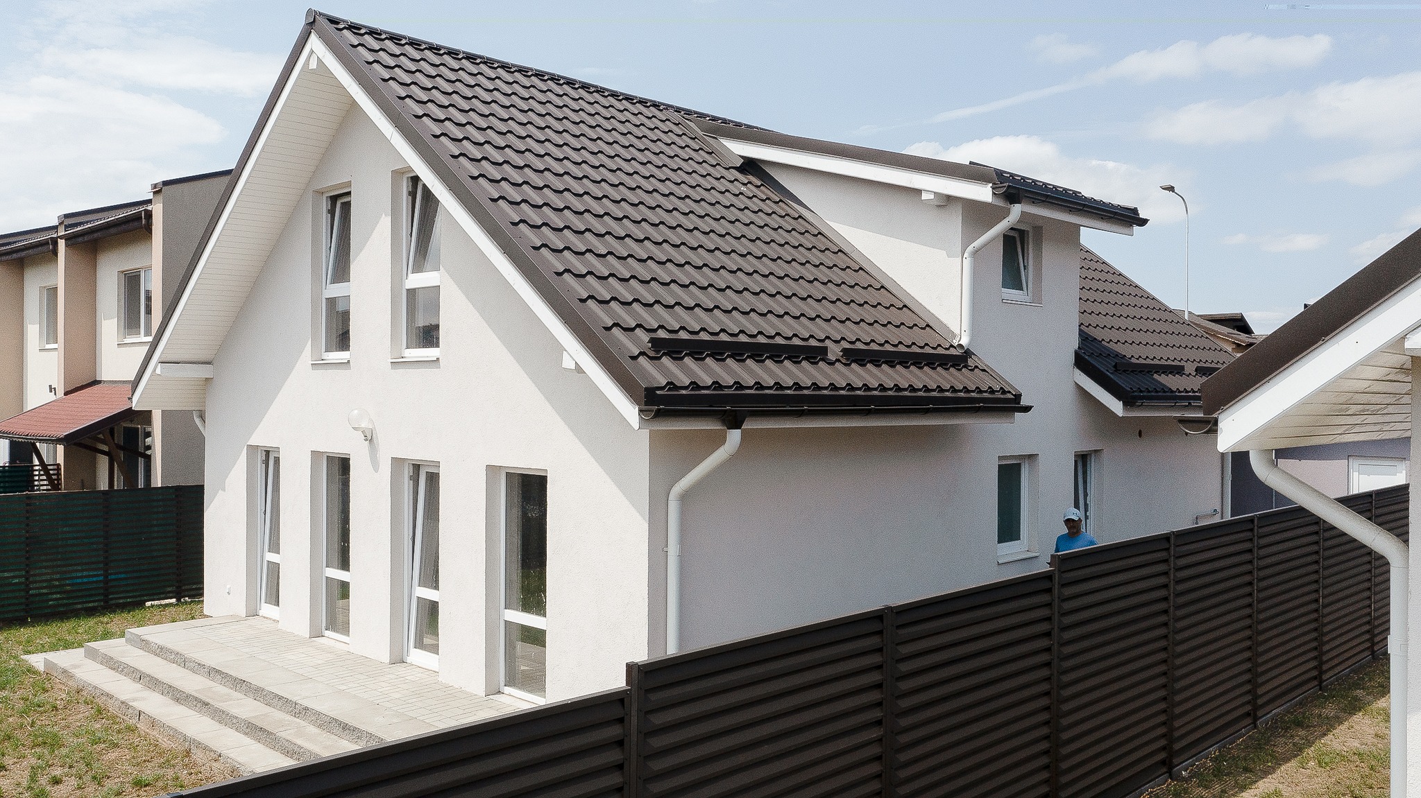 O casă modernă cu două etaje, cu acoperiș din țiglă închisă la culoare și pereți albi. Dispune de ferestre mari și o curte împrejmuită, sub un cer albastru senin, emanând o atmosferă contemporană și primitoare.