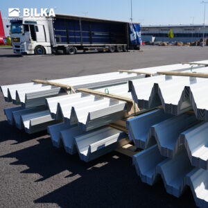 Stive de tablă albă de acoperiș metalică pe paleți de lemn, cu un camion cu laterale deschise în fundal.