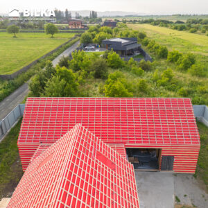 Vedere aeriană a unei case vibrante cu acoperiș roșu, situată în mijlocul unei vegetații luxuriante și a unor câmpuri. Peisajul prezintă un amestec de clădiri moderne și rustice.