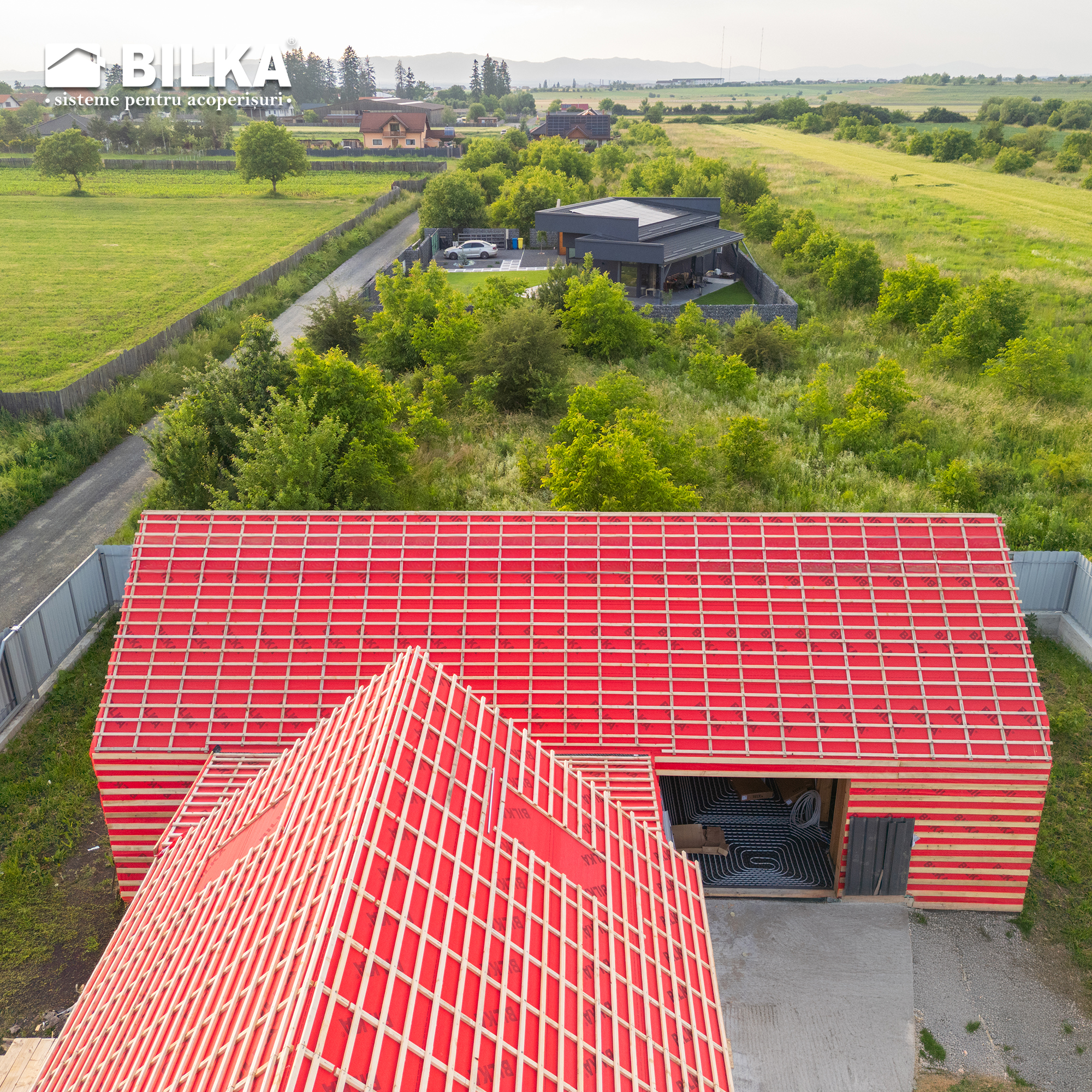 Vedere aeriană a unei case vibrante cu acoperiș roșu, situată în mijlocul unei vegetații luxuriante și a unor câmpuri. Peisajul prezintă un amestec de clădiri moderne și rustice.