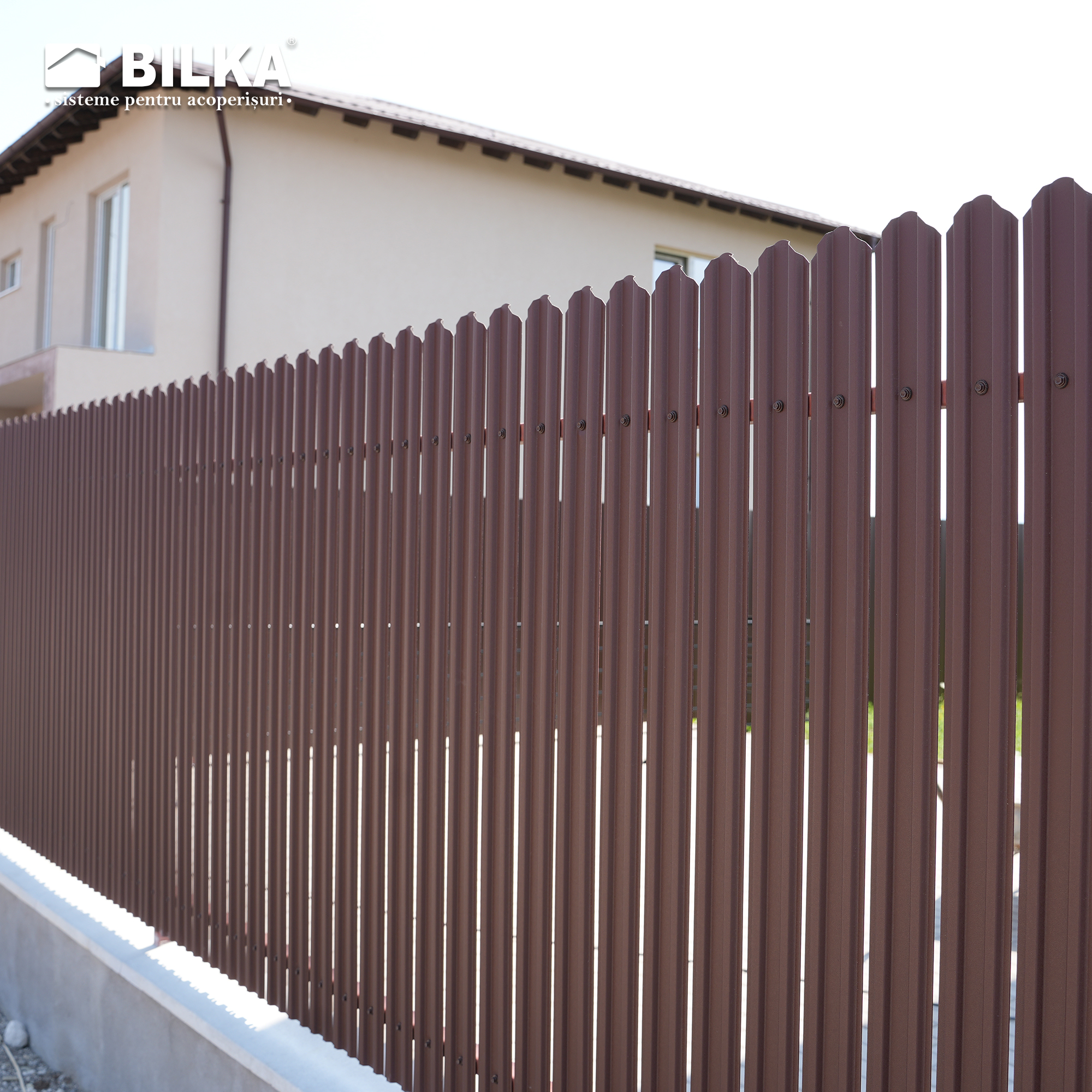 Un gard metalic maro cu panouri verticale se află în fața unei case de culoare crem cu acoperiș înclinat. Scena transmite intimitate și siguranță.