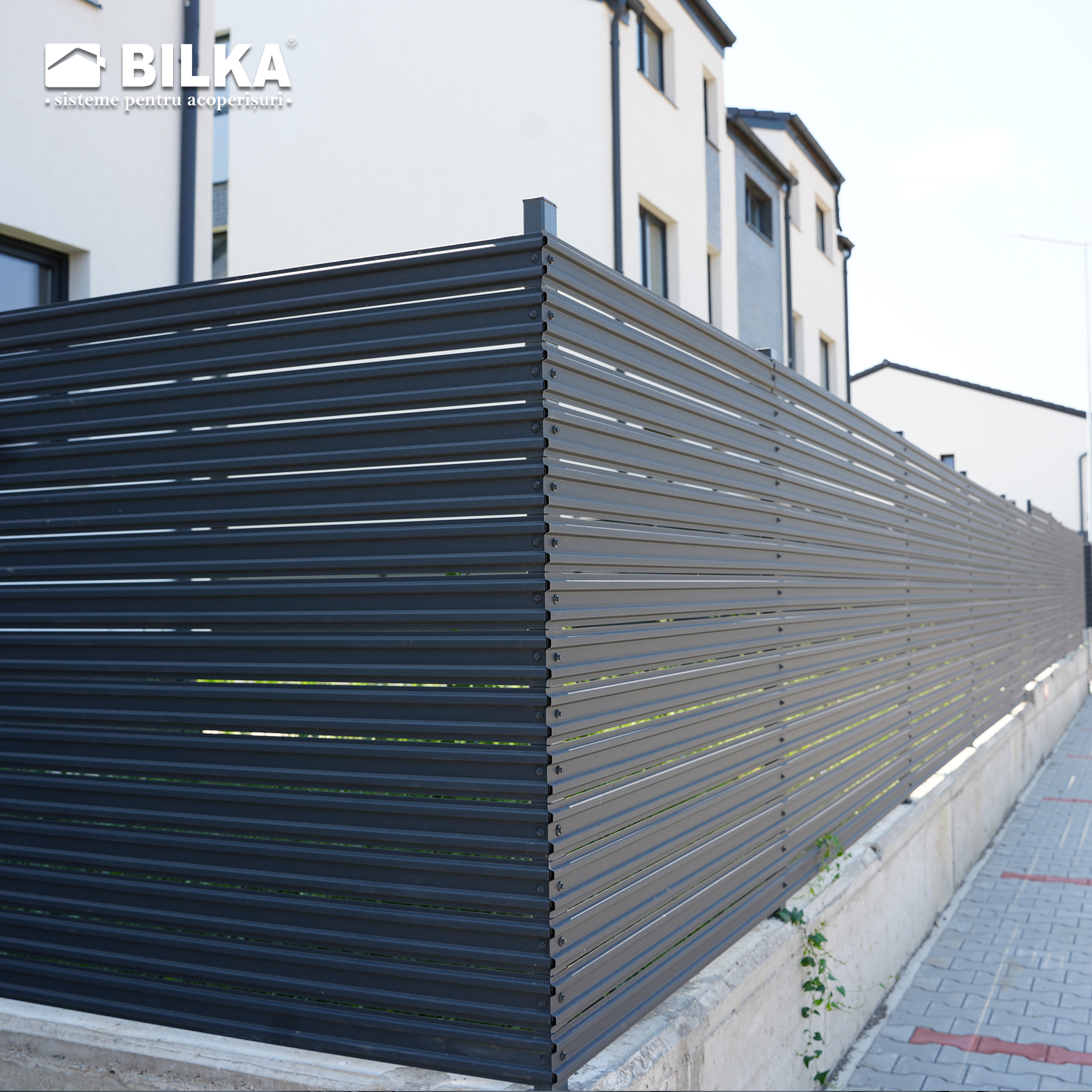 Un gard metalic orizontal modern, gri închis, mărginește un trotuar lângă o clădire bej, transmițând siguranță și design contemporan.