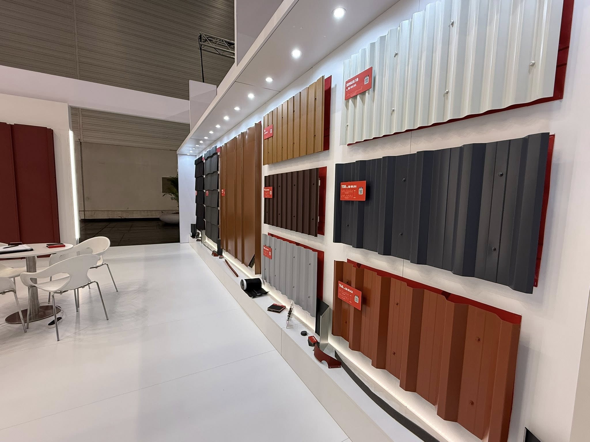 Stand expozițional care expunea pe un perete luminat diverse foi metalice de acoperiș în diferite culori și texturi. În apropiere, o masă cu scaune albe.