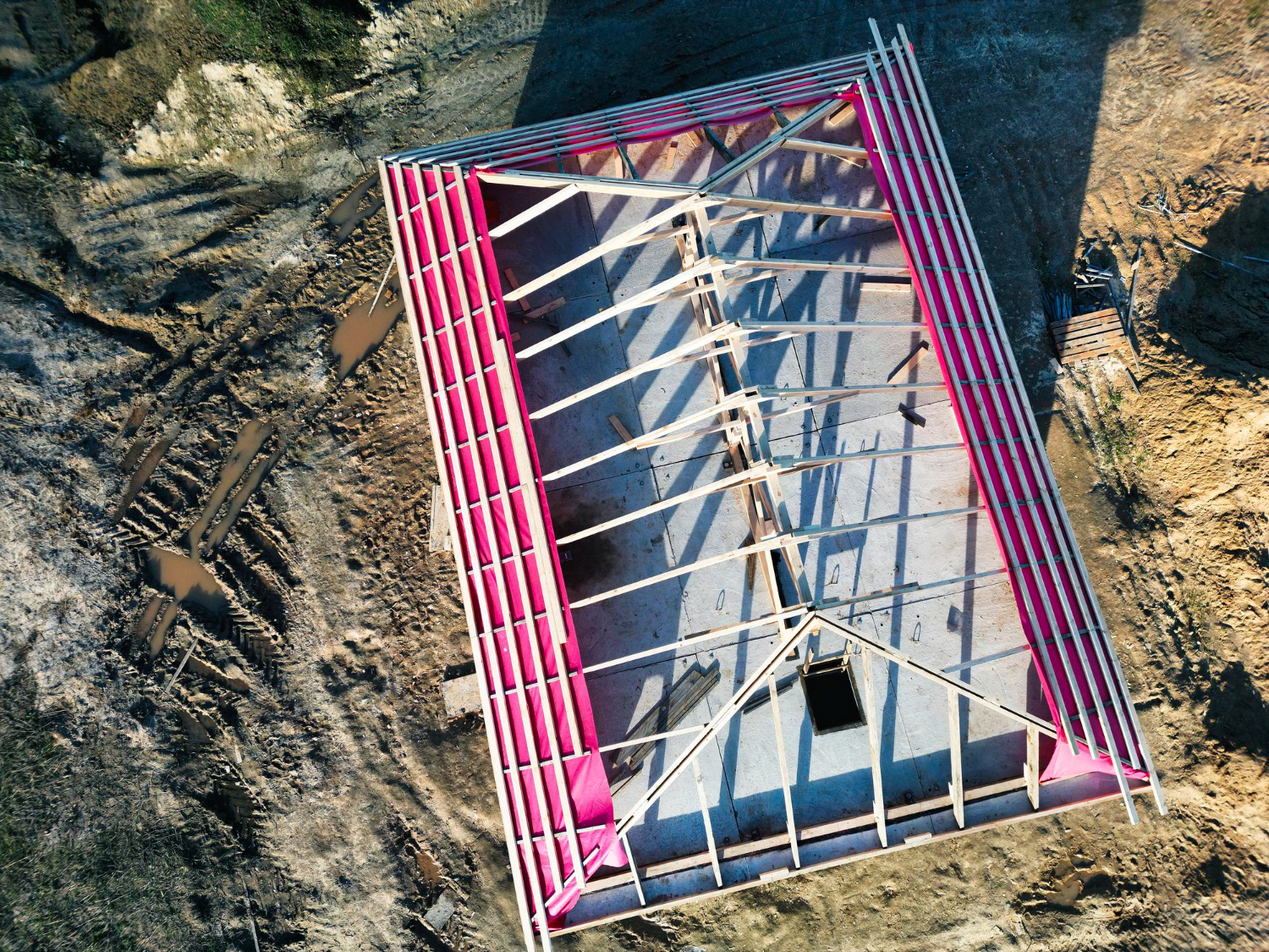 Vedere aeriană a unei case în construcție, cu grinzi de lemn și izolație roz care căptușește acoperișul