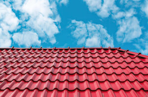 Acoperiș metalic roșu cu țigle ondulate sub un cer albastru strălucitor, brăzdat de nori albi, transmițând un sentiment de liniște și eficiență.