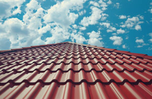 Acoperiș roșu din tablă ondulată care se înalță spre un cer albastru strălucitor, presărat cu nori albi pufoși, transmițând un sentiment de deschidere și stabilitate.