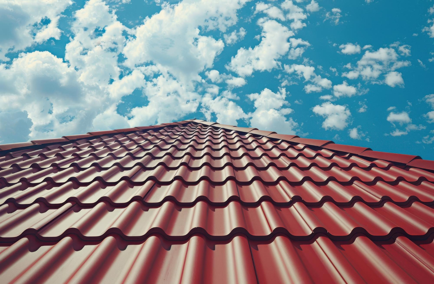 Acoperiș roșu din tablă ondulată care se înalță spre un cer albastru strălucitor, presărat cu nori albi pufoși, transmițând un sentiment de deschidere și stabilitate.