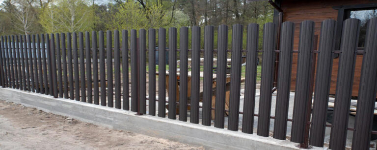 O gardă metalică cu lamele verticale, de culoare maro închis, așezată pe o bază de beton, delimitează o curte. În fundal se văd copaci cu frunziș de primăvară și o clădire din lemn.