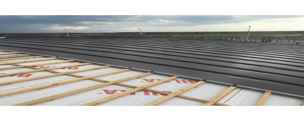 Un acoperiș metalic de culoare închisă, montat parțial peste o structură formată din șipci de lemn și plăci izolante. Cerul este înnorat, ceea ce lasă să se întrevadă o furtună iminentă.