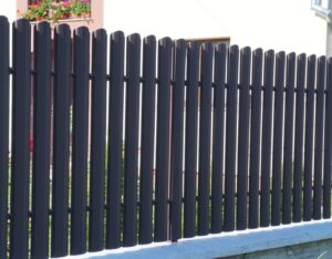 Un gard din metal negru cu lamele verticale, luminat de razele soarelui. În fundal se vede o casă cu jardiniere și flori roz, creând o imagine îngrijită și plină de viață.