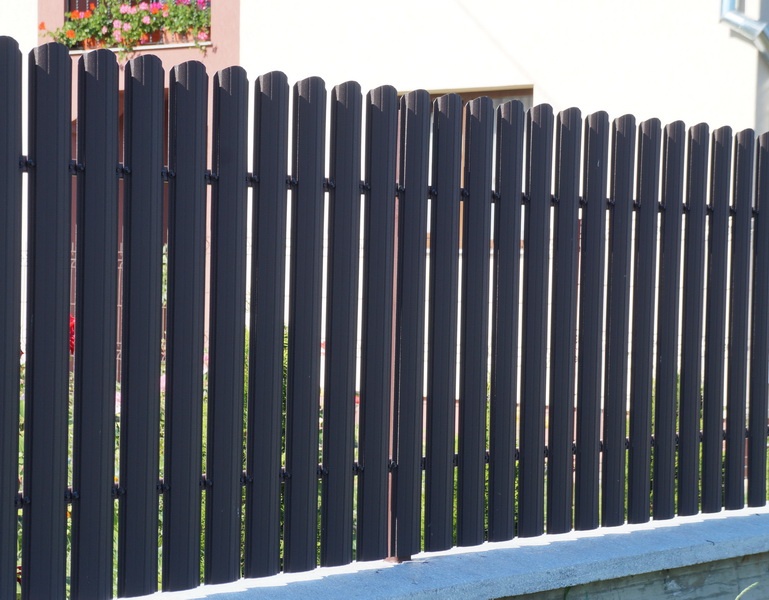 Un gard din metal negru cu lamele verticale, luminat de razele soarelui. În fundal se vede o casă cu jardiniere și flori roz, creând o imagine îngrijită și plină de viață.