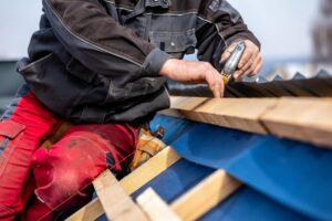 O persoană îmbrăcată într-o jachetă închisă la culoare și pantaloni roșii lucrează pe un acoperiș albastru, aliniind cu precizie grinzile de lemn cu ajutorul unei unelte. Scena transmite o senzație de concentrare.